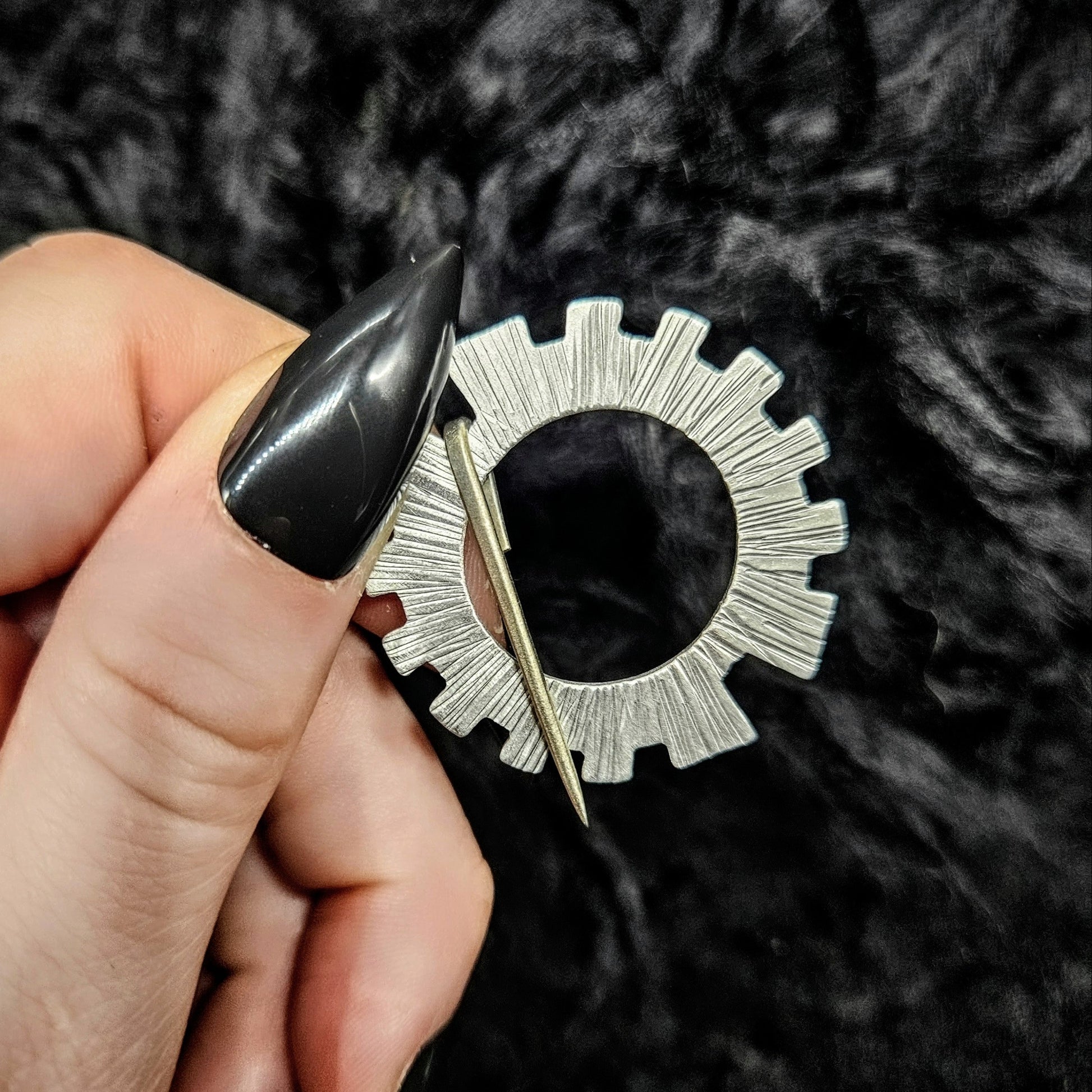 Hand holding a silver gear shaped brooch pin with a black background