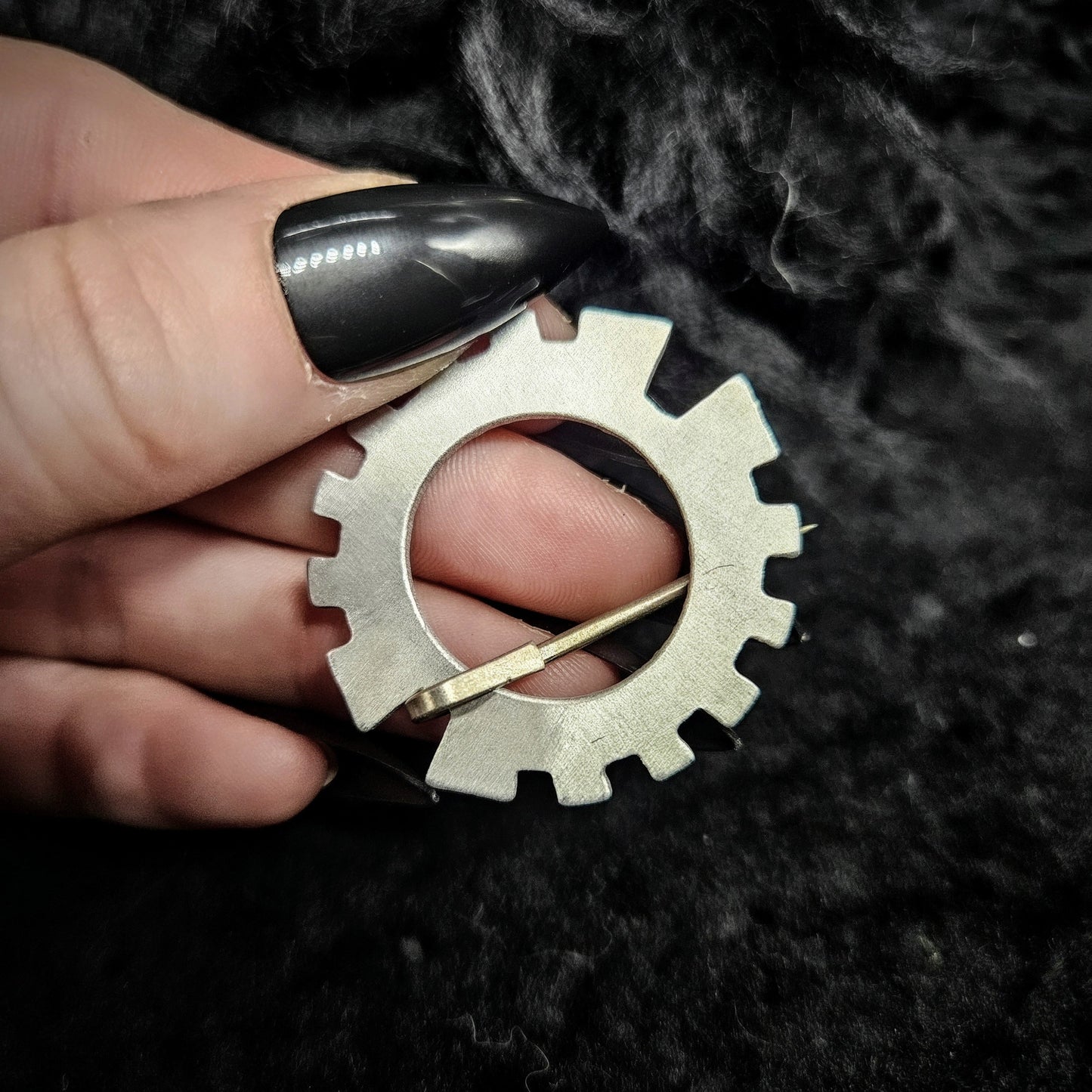 Metal gear-shaped earring held by a person with black nail polish on a dark background