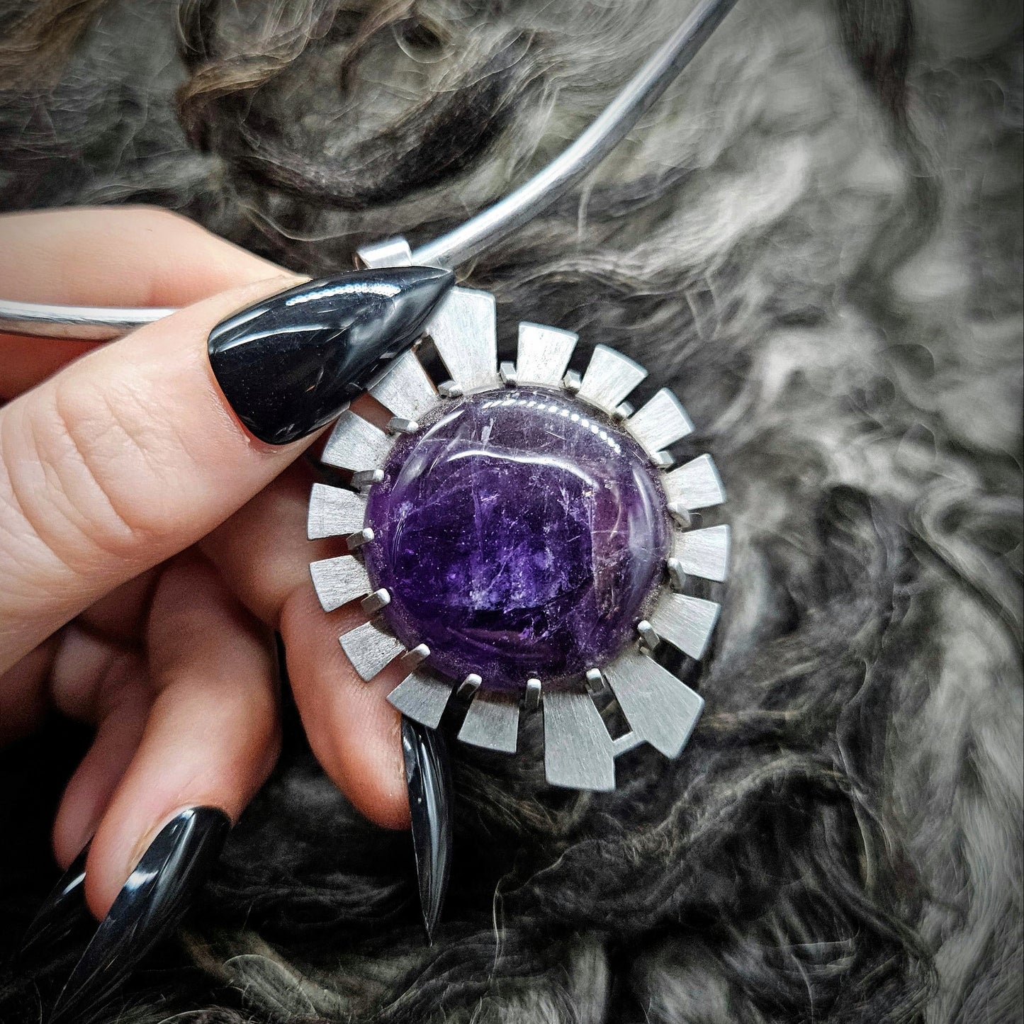 Hand holding a silver and purple gemstone pendant against a textured fur background