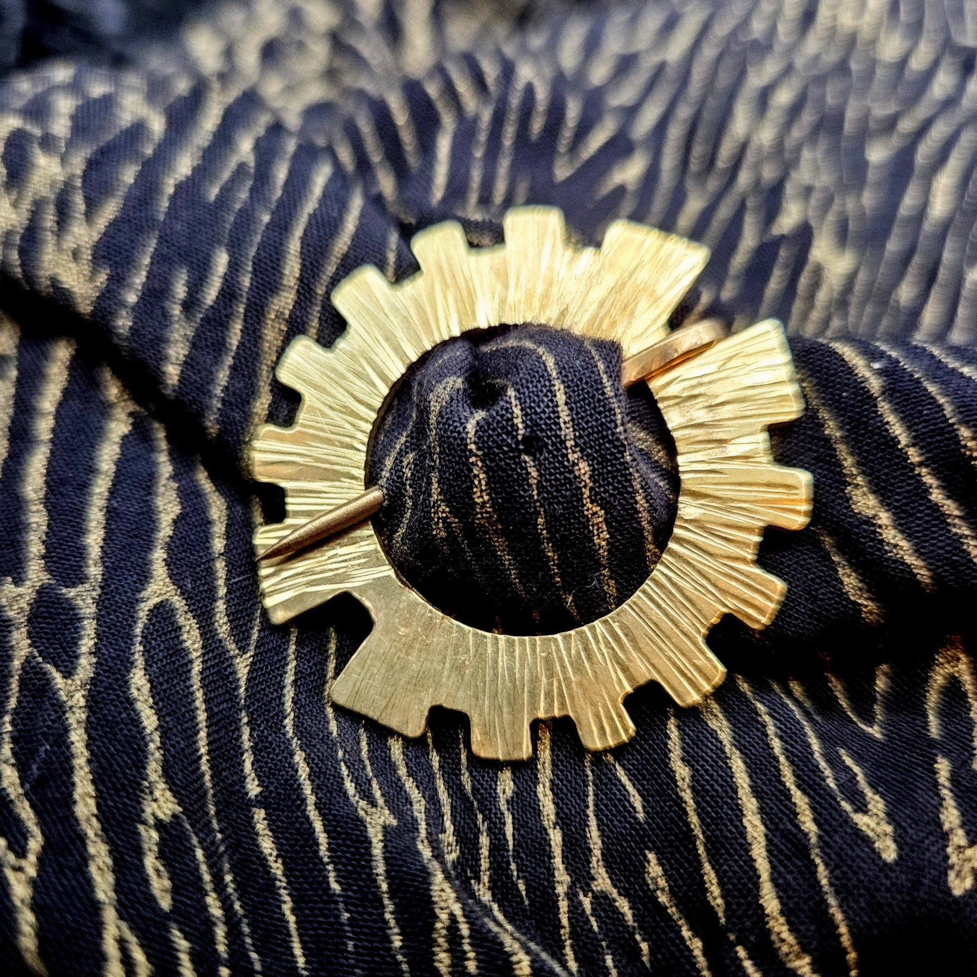 Gold gear shaped brooch pinned on a  black and gold fabric scarf.